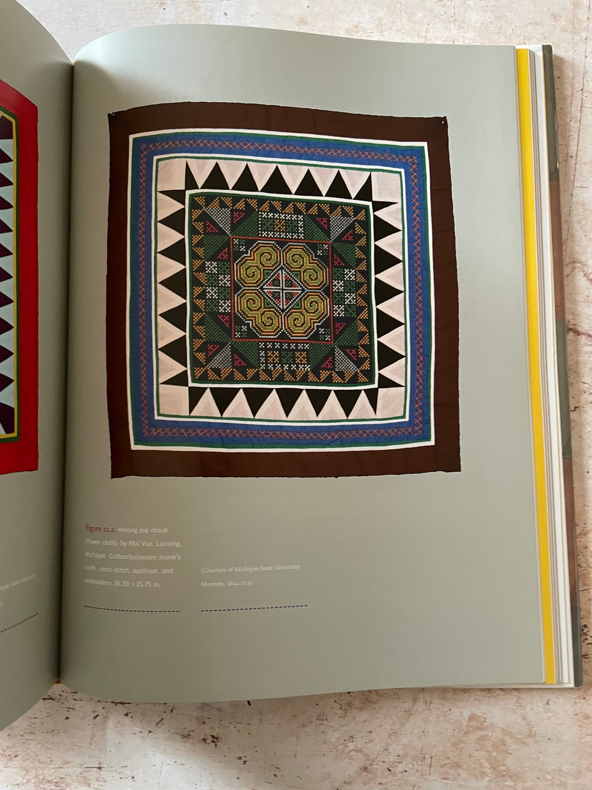 An open book  page showing a colorful geometric textile  centered on a muted sage-green page, photographed from above on a light stone-like surface.