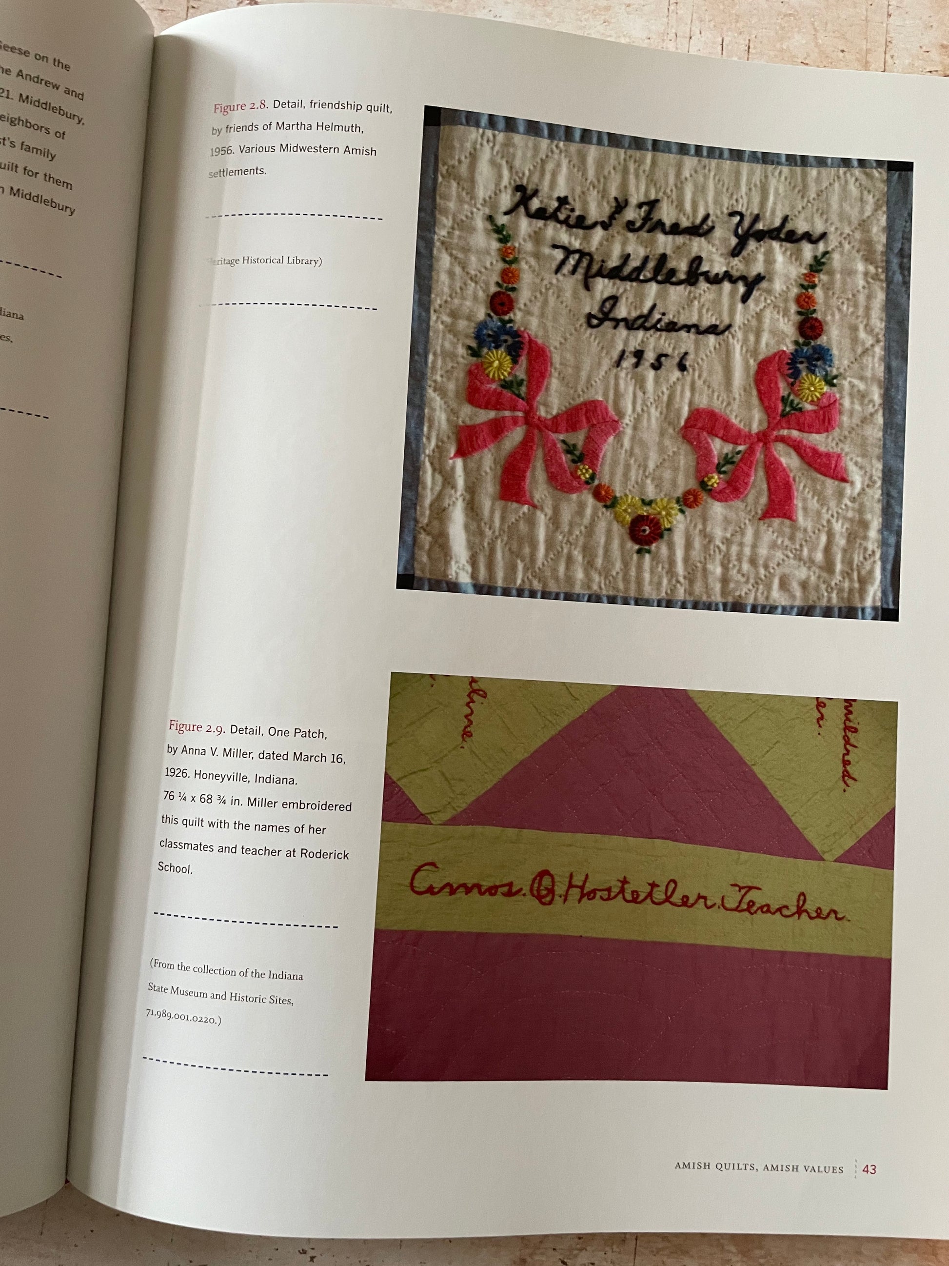 An open book showing printed pages with two quilt photos and handwritten notes. The top photo shows a quilt with embroidered bows and  text, and the bottom photo shows a quilt with geometric sections.