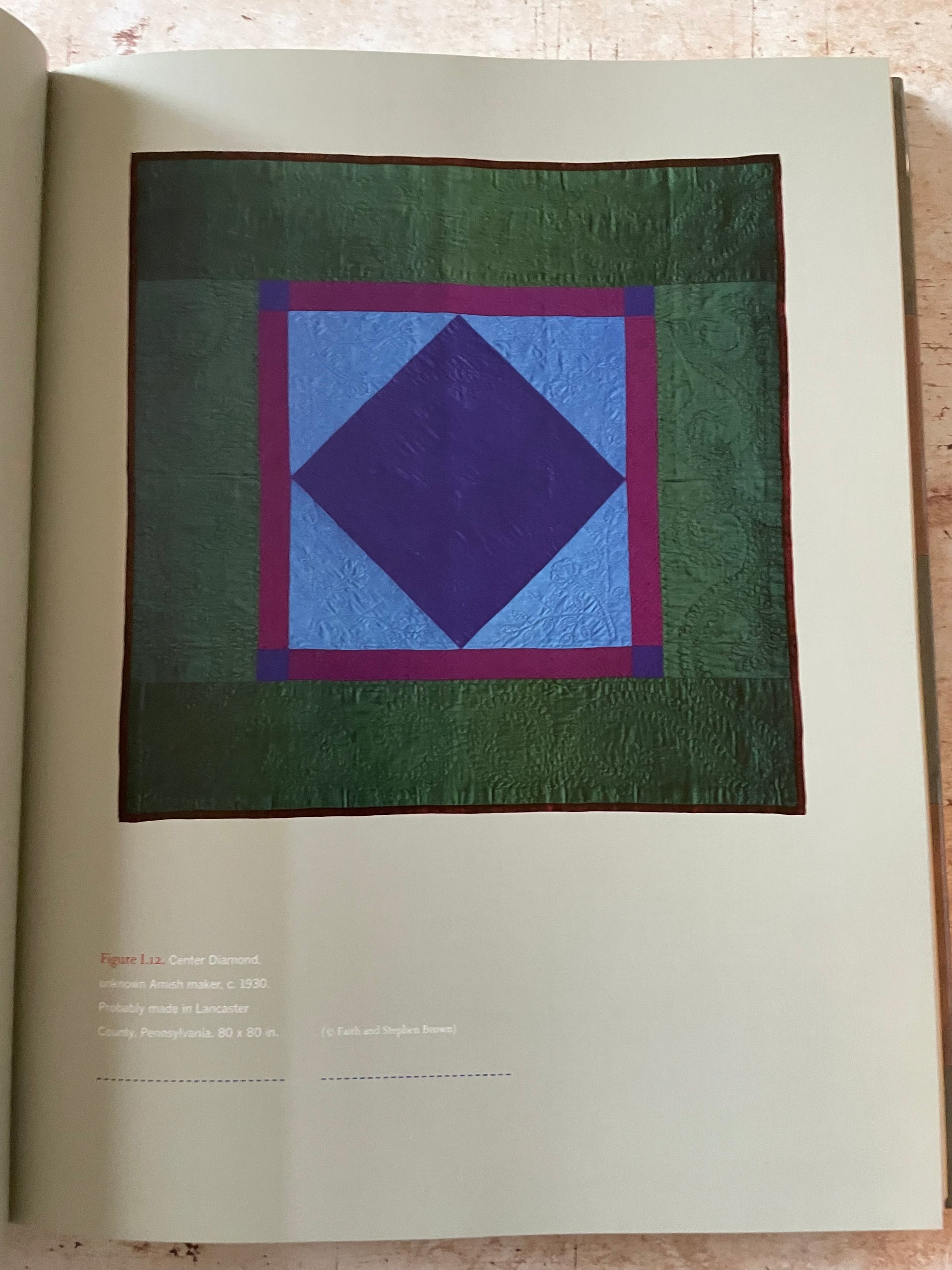 A quilt displayed on a book page, featuring a geometric pattern with concentric squares in dark green, purple, light blue, and magenta. 