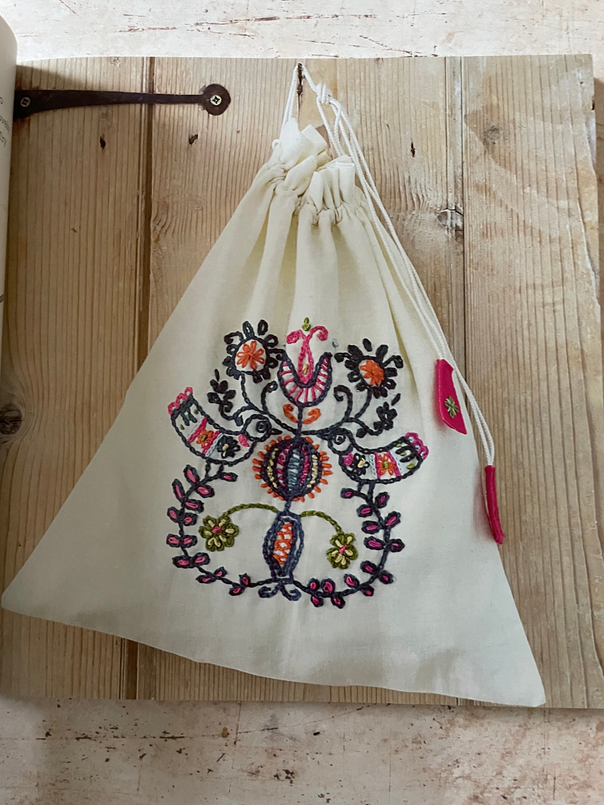 An off-white drawstring bag with a colorful embroidered floral and paisley pattern, resting on a rustic wooden plank background.