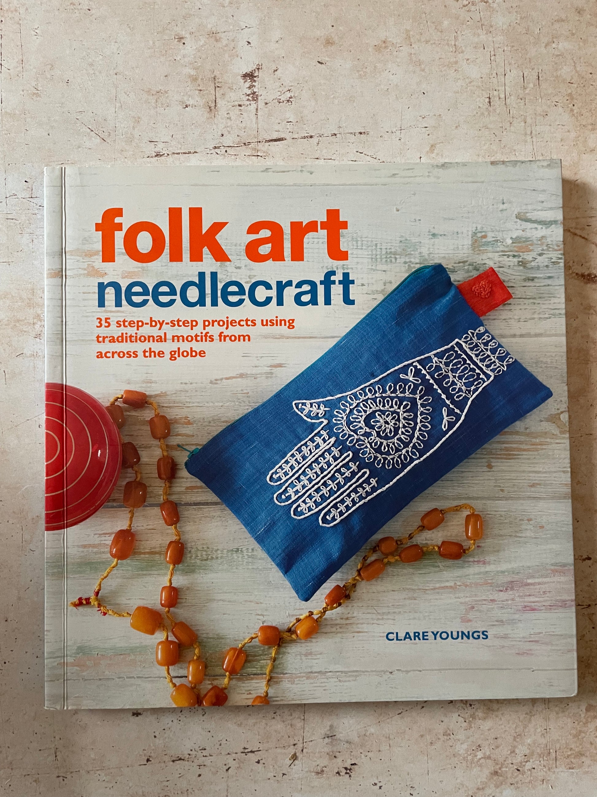 The front cover of the book 'folk art needlecraft' by Clare Youngs, featuring a blue embroidered hamsa hand pouch and amber beads on a whitewashed wood background.