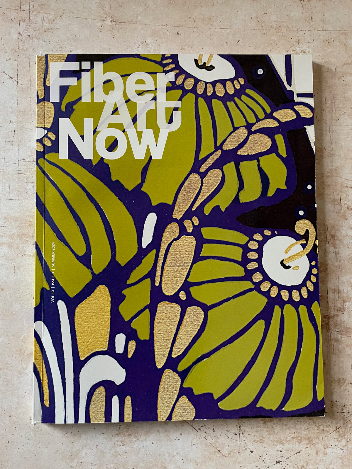 A book or magazine titled 'Fiber Art Now' with a bold abstract cover design in olive green, navy blue, white, and gold, photographed standing against a light beige textured wall.