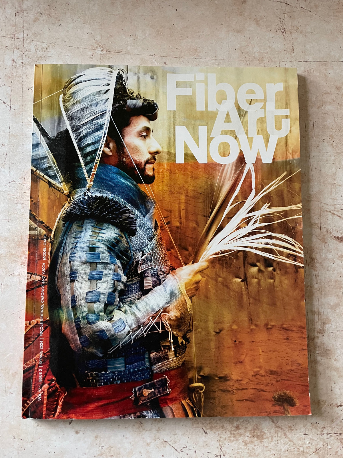 A physical copy of the 'Fiber Art Now' publication or book cover, featuring a painted illustration of a weaver in traditional attire holding fiber, displayed flat on a textured concrete surface.