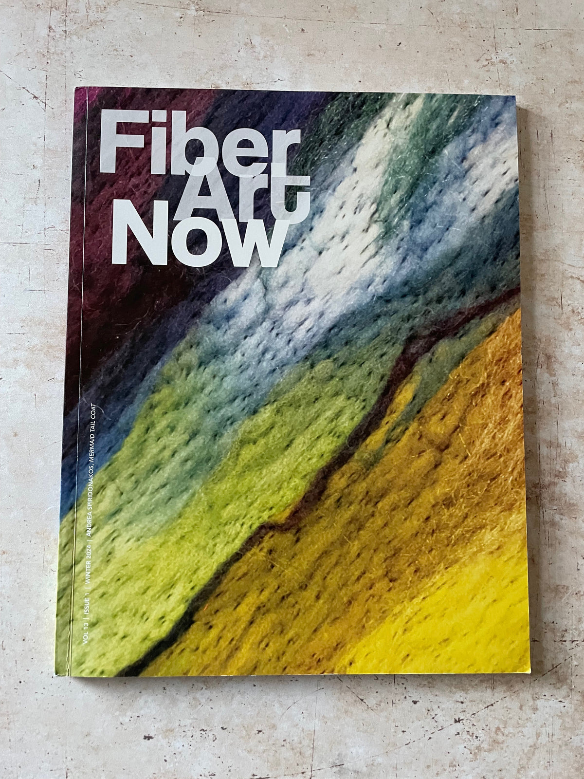 A physical copy of the 'Fiber Art Now' publication, featuring a colorful abstract cover design with the title in bold white text, photographed flat on a textured beige stone surface.