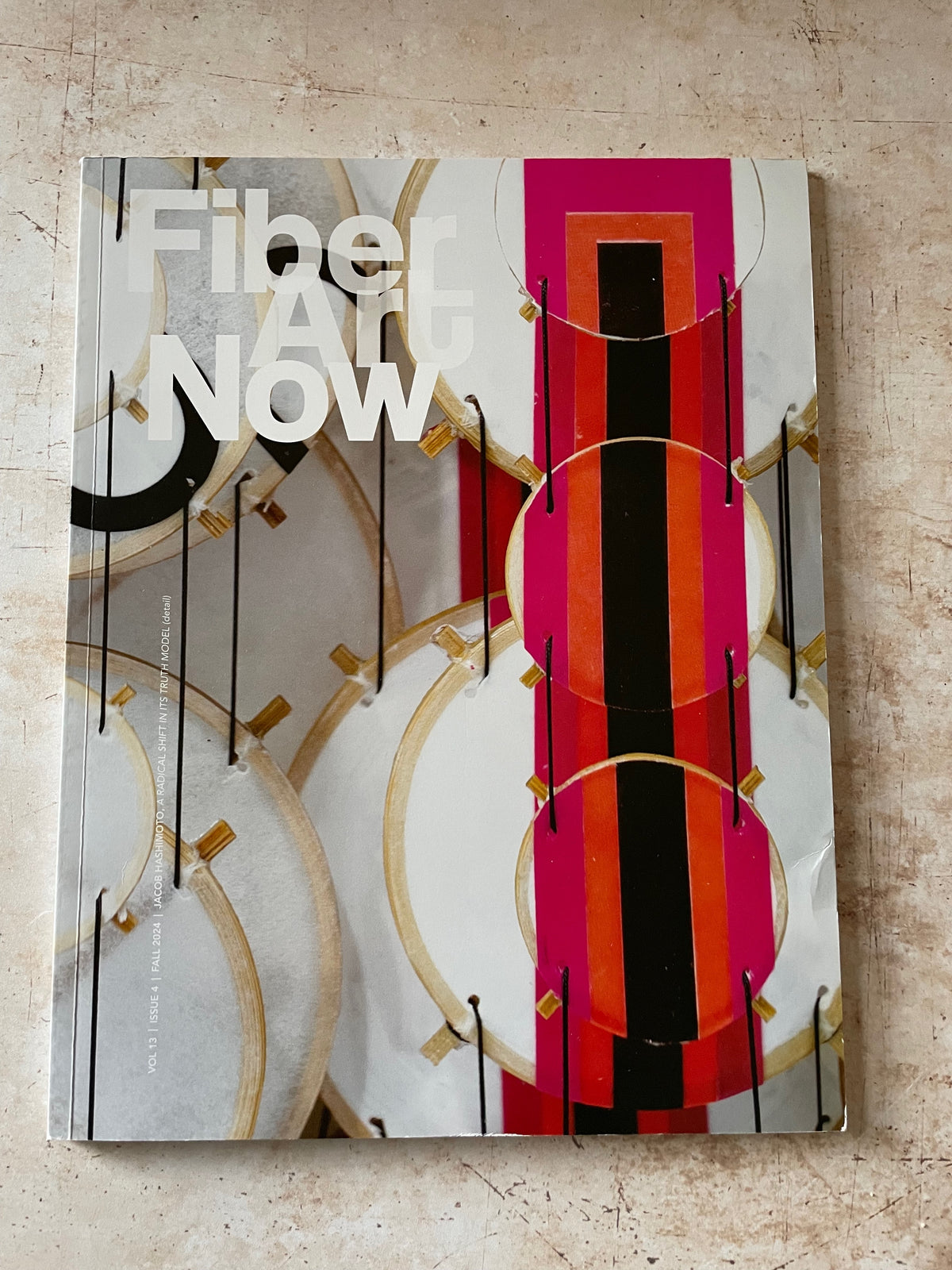 A magazine titled "Fiber Art Now" lying on a textured beige surface. The cover features an abstract geometric design with white, black, pink, orange, and gold elements.