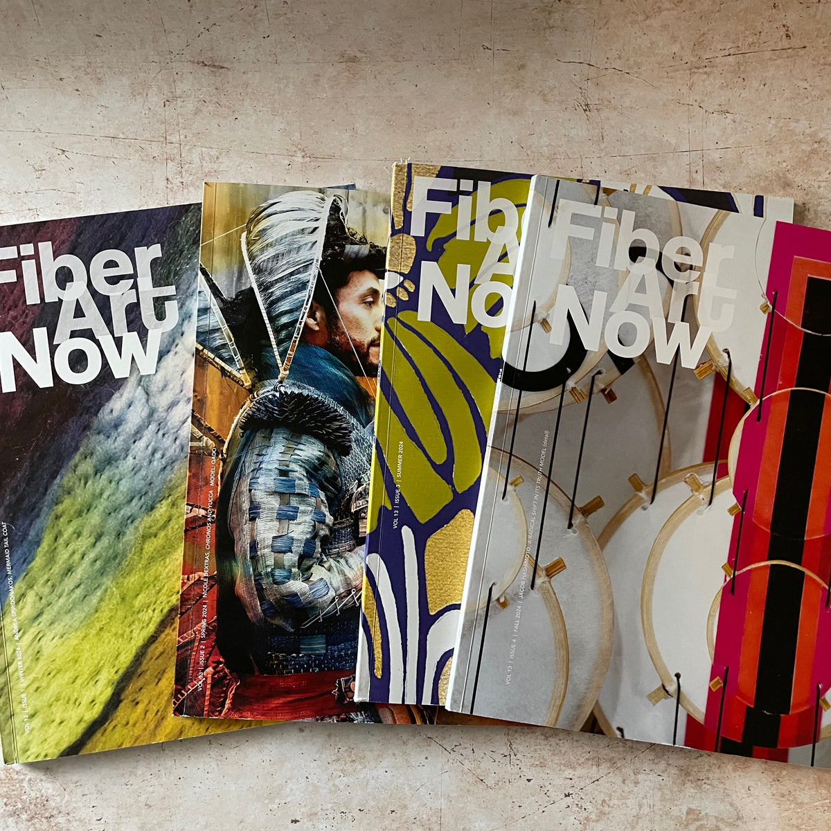A fan of printed art magazines titled 'Fiber Art Now' spread out on a textured beige surface. The covers feature diverse colorful artistic designs, including abstract landscapes, a figure in armor, and geometric patterns.