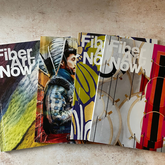 A fan of printed art magazines titled 'Fiber Art Now' spread out on a textured beige surface. The covers feature diverse colorful artistic designs, including abstract landscapes, a figure in armor, and geometric patterns.