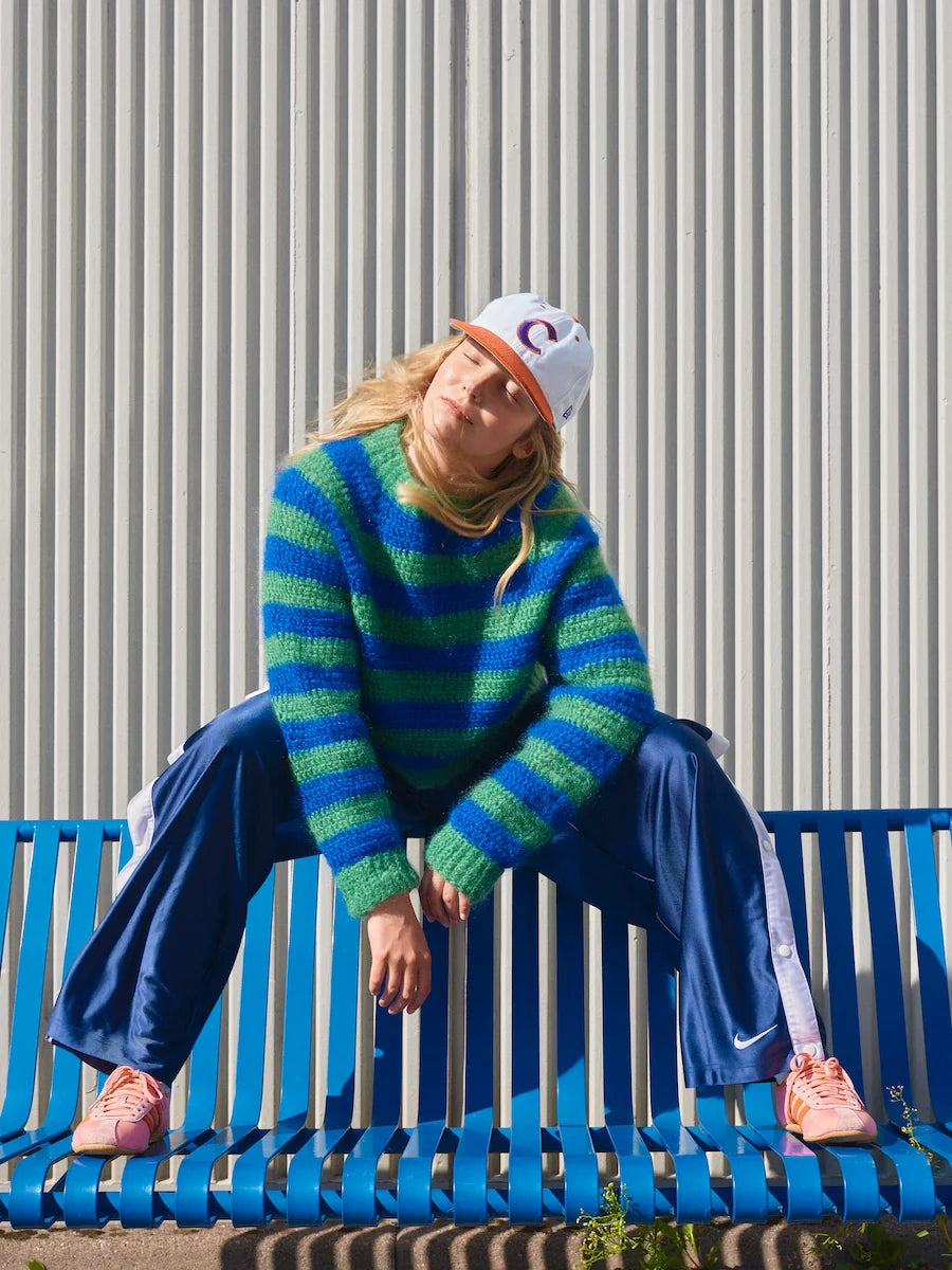 Person wearing a blue and green striped sweater and blue pants sitting on a blue bench against a gray wall.