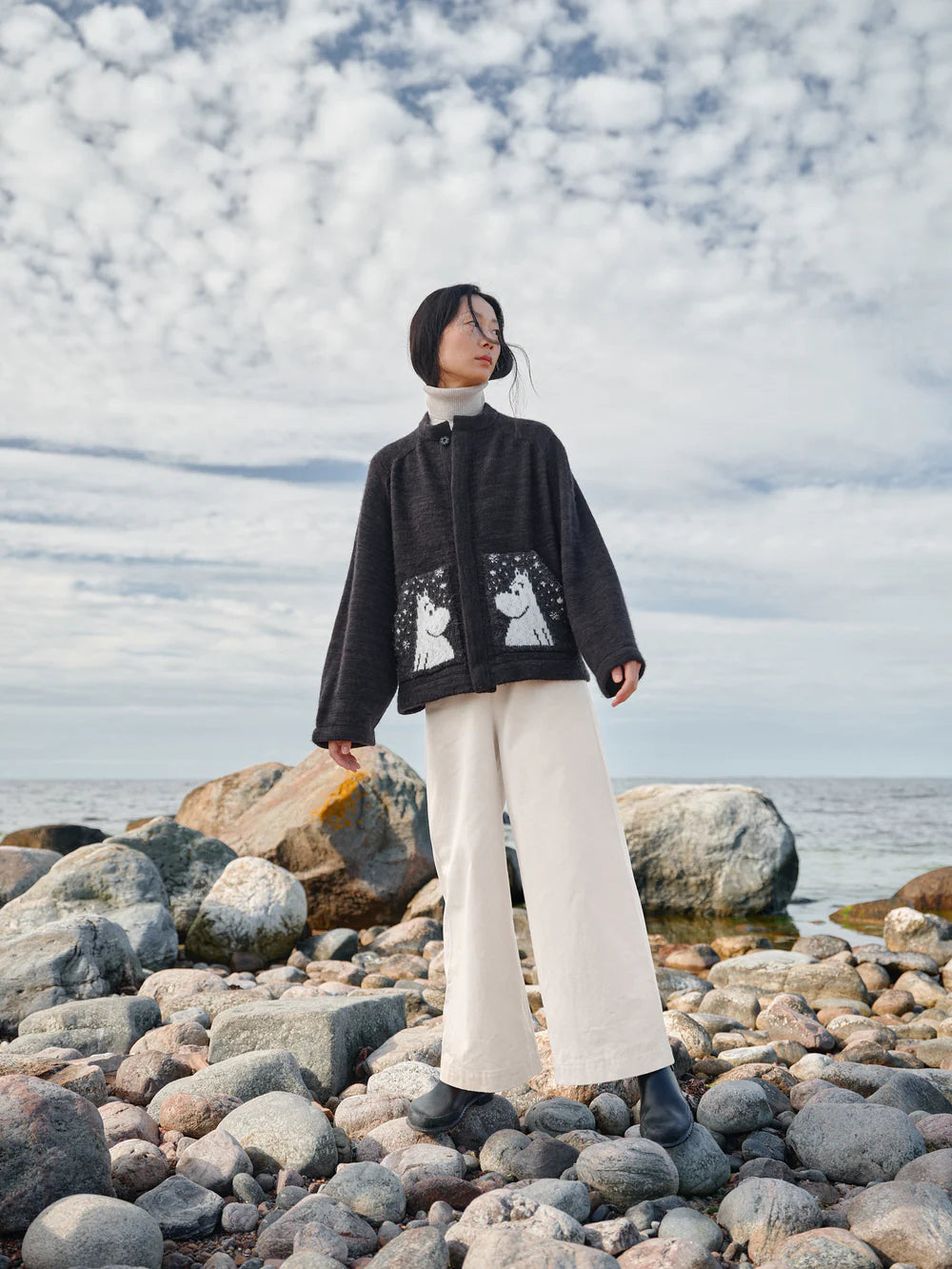 Person standing on a rocky beach with a black jacket and white pants, holding a yellow object.