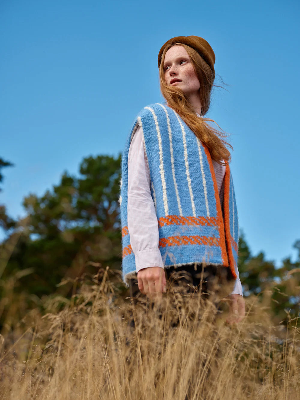 Woman wearing a colorful striped vest and brown beret standing in a field with a clear blue sky.