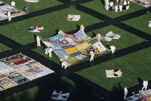 An aerial view shows several people standing on a green lawn while handling large colorful printed fabric panels laid out in a grid-like arrangement.