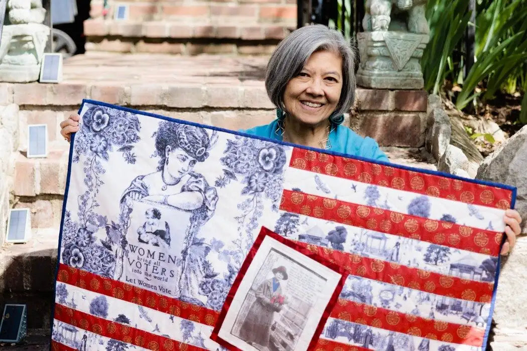 A smiling woman with gray hair holds up a large quilted American flag design featuring blue-and-white historical illustrations and red-and-white stripes, with a small inset photo attached near the lower center.