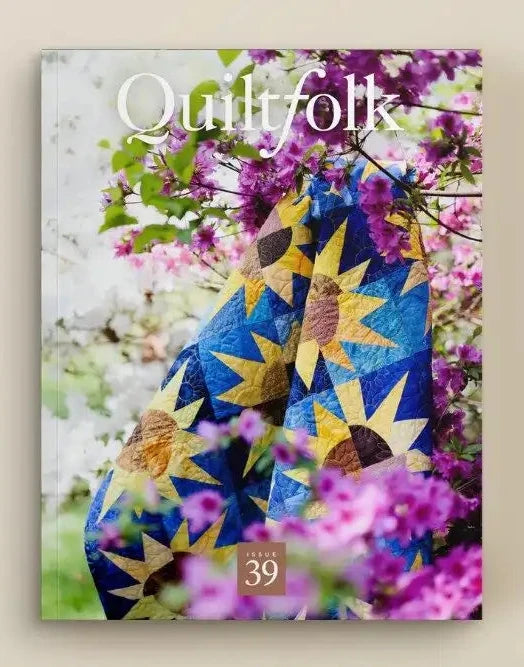 A quilted fabric panel with a blue and yellow starburst pattern, draped over a floral quilted surface with purple flowers and the text 'Quilt folk' visible, set against a plain beige background.