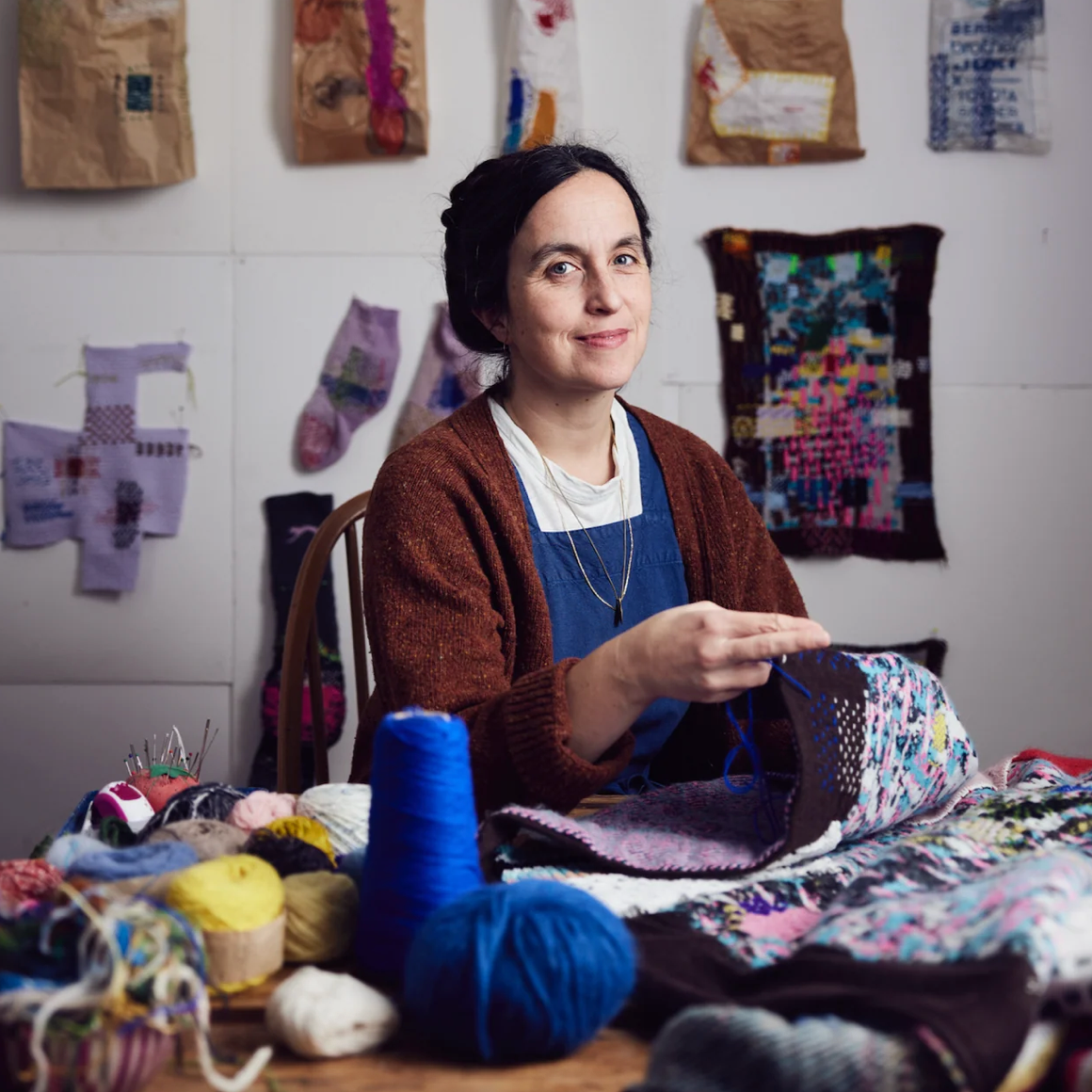 A woman sits at a table surrounded by colorful balls of yarn and knitted fabric, smiling as she works on a textile piece. The background shows a white wall decorated with handmade quilts and fabric samples.