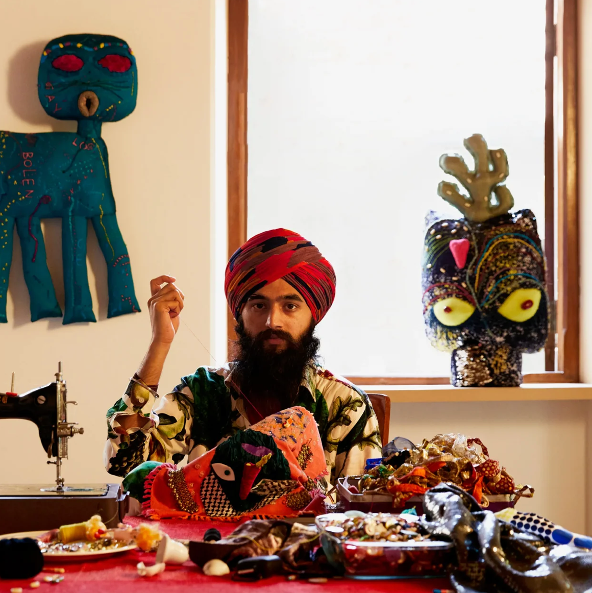 A man with a beard and a red patterned turban sits at a table covered with colorful handmade textiles and objects, surrounded by a bright window and decorative wall pieces.