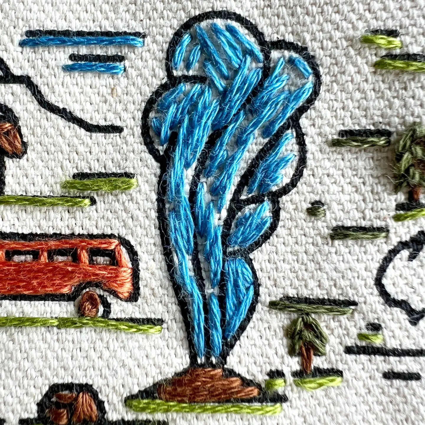 Embroidery design of a blue tree with a bus and people in the background on white fabric.