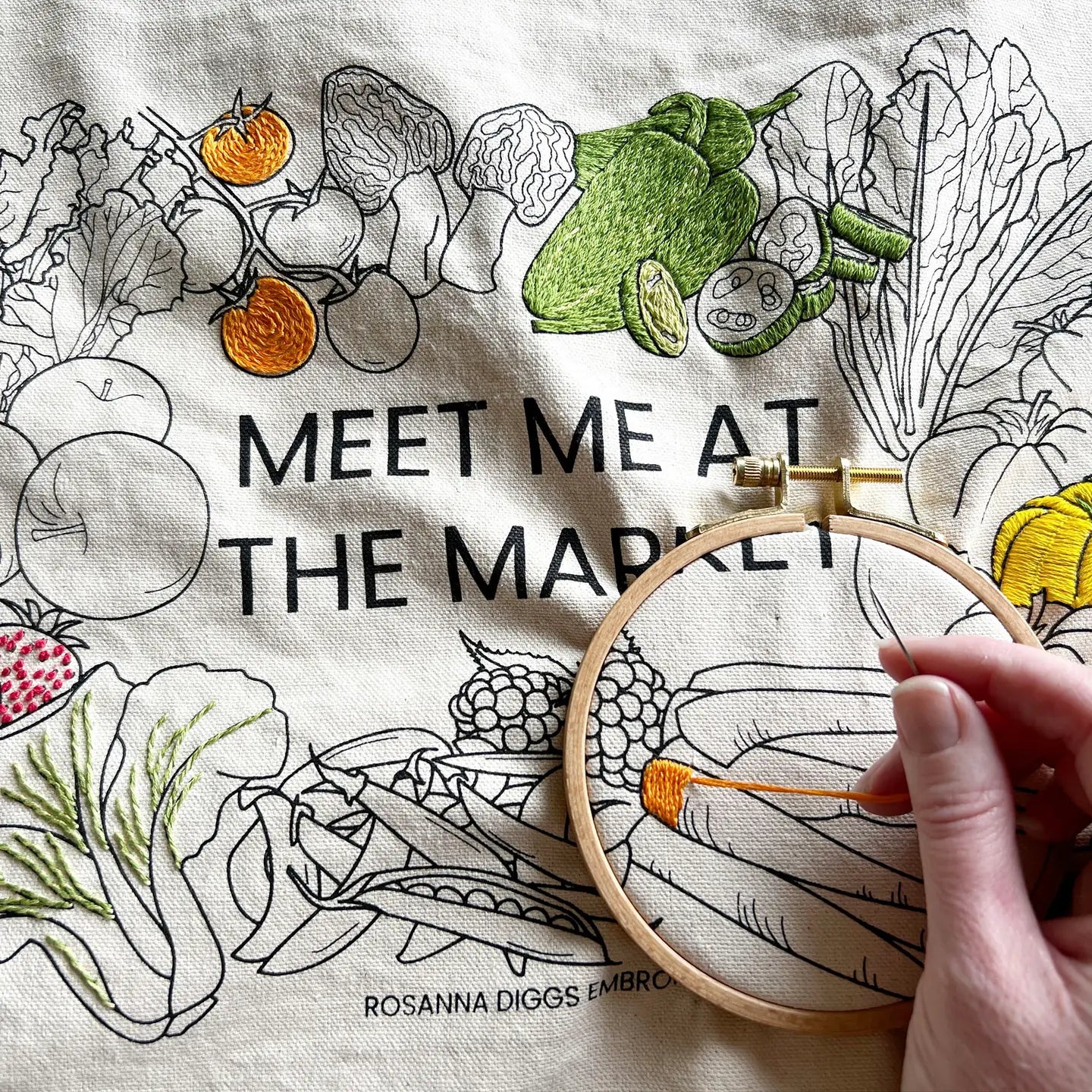 Embroidery hoop with embroidered vegetables and text 'Meet Me at the Market' on fabric.