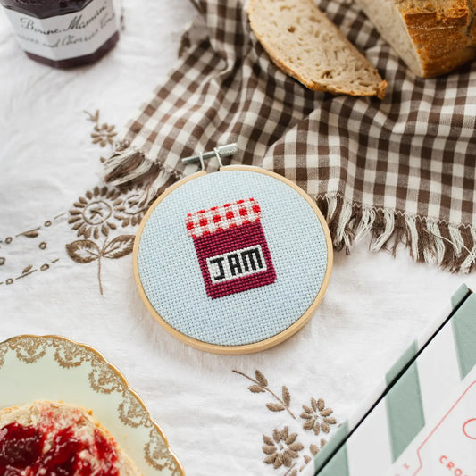 Cross-stitched design of a jam jar on a table with bread and jam.