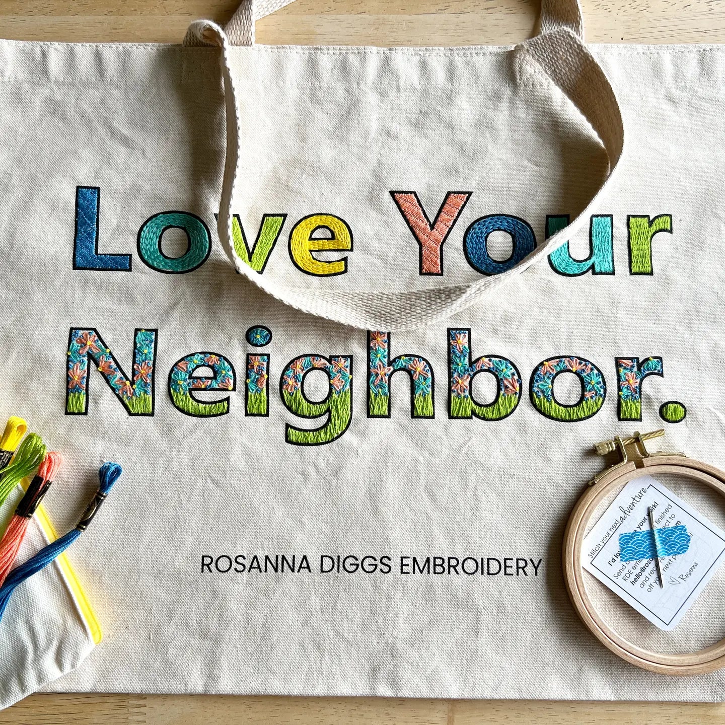 Tote bag with 'Love Your Neighbor' text and embroidery hoop on a wooden surface, featuring Rosanna Diggs Embroidery.