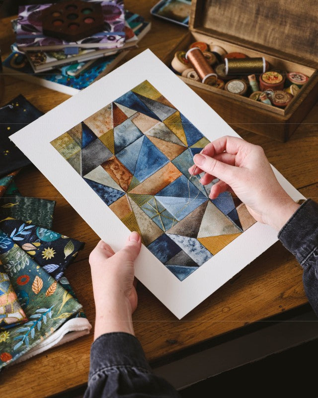 Person stitching on a watercolor painted quilt with sewing tools in the background