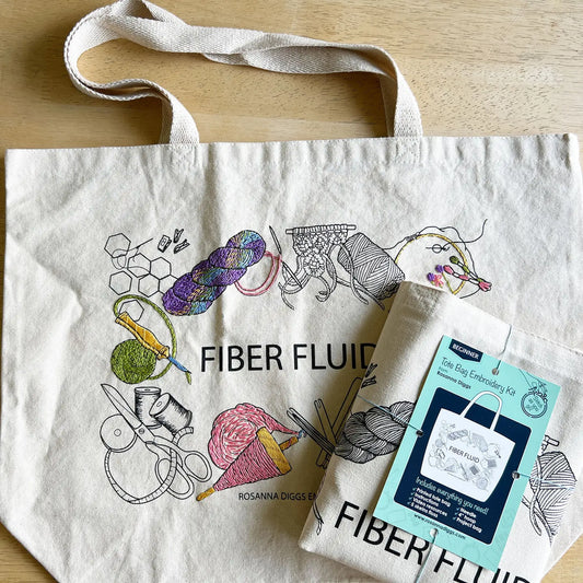 White tote bag with fiber-themed illustrations and a tag labeled 'Fiber Fluid' on a wooden surface.
