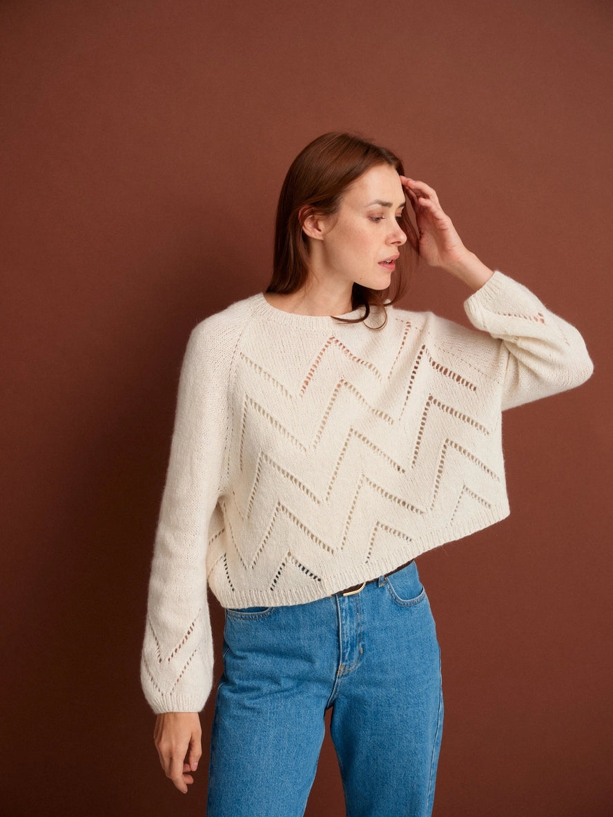 Woman wearing a white patterned sweater and blue jeans against a brown background