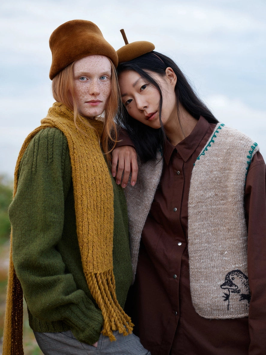 Two people wearing autumn-themed clothing with scarves and hats against a natural background.