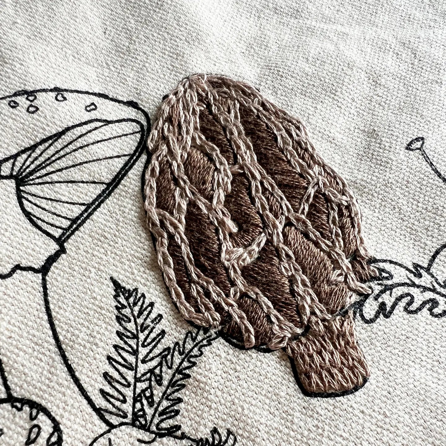 Embroidered brown mushroom on a light fabric background with black line art of plants and leaves.