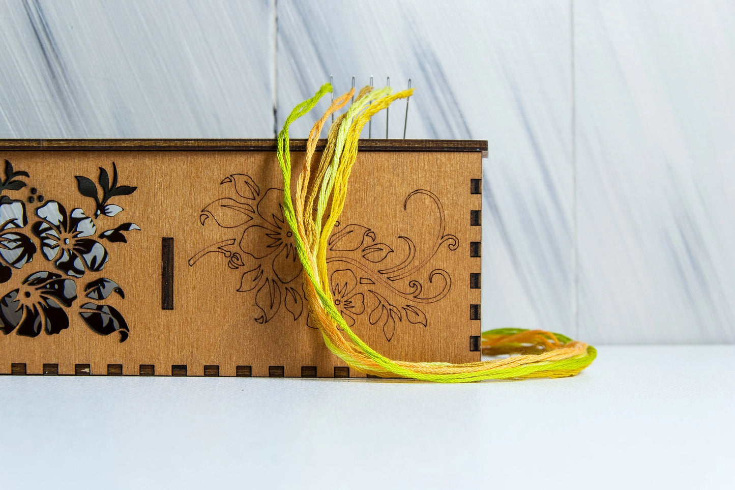 Wooden box with floral designs and a yellow threaded needle on a white surface.
