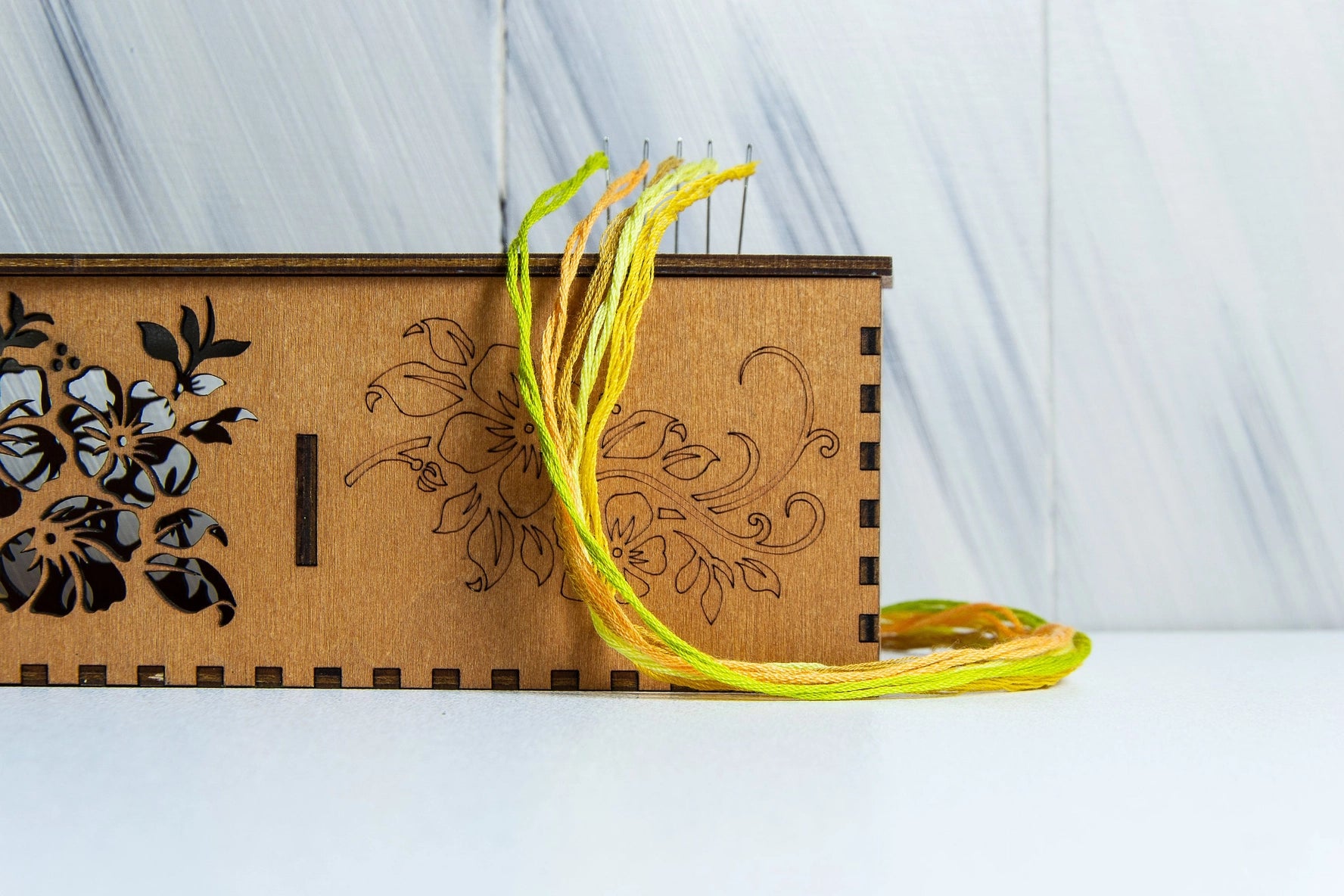 Wooden box with floral designs and a yellow threaded needle on a white surface.