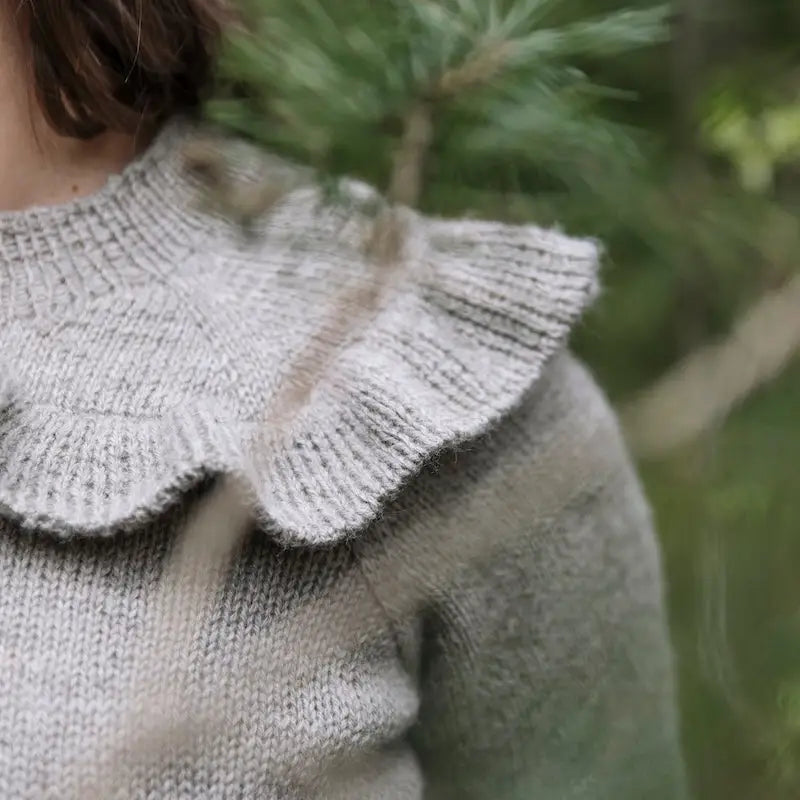 Close-up of a person wearing a knitted sweater with a ruffled collar against a blurred natural background.
