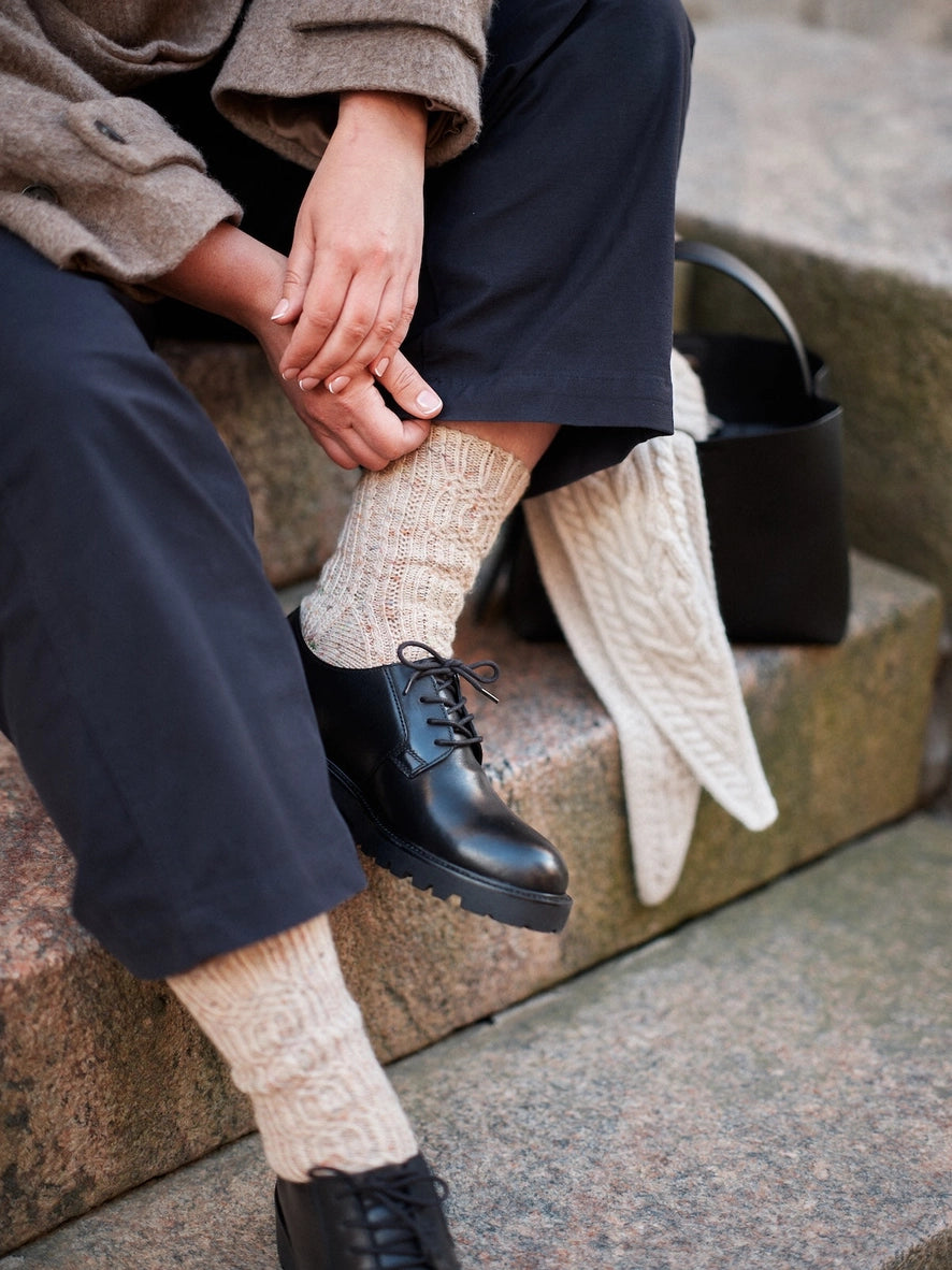 A person sitting on stone steps wearing dark navy trousers, cream cable-knit socks, black lace-up boots, and a taupe coat. A black handbag rests beside them on the steps.